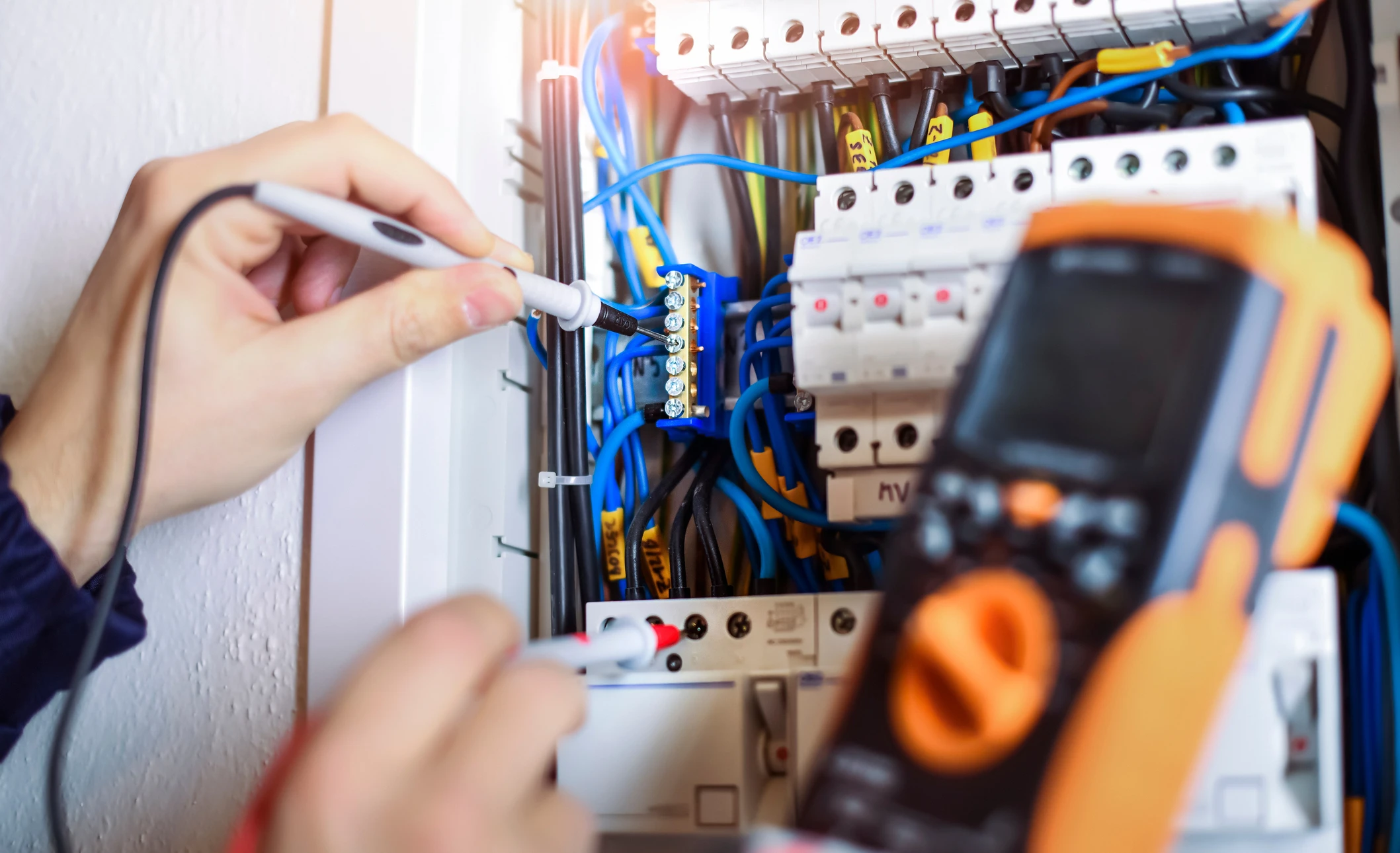 Un électricien travaille sur un tableau électrique domestique, utilisant un multimètre orange pour effectuer des mesures